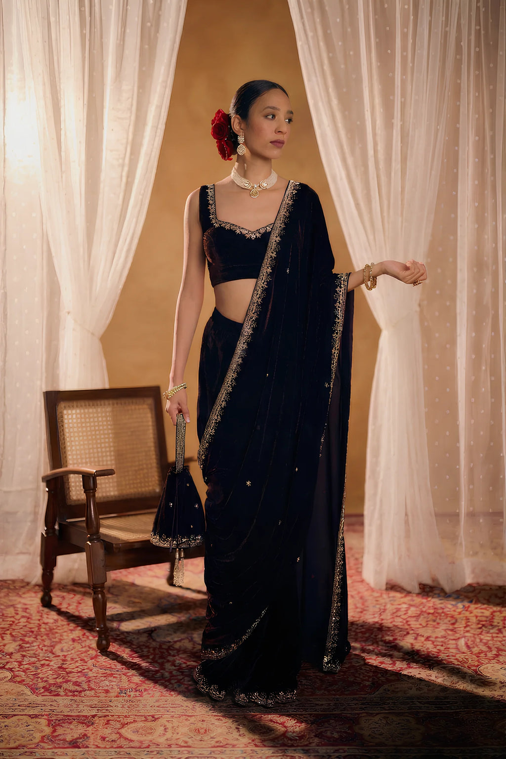 Woman in a black valvat saree with gold details standing in a room with white curtains and a wooden chair.