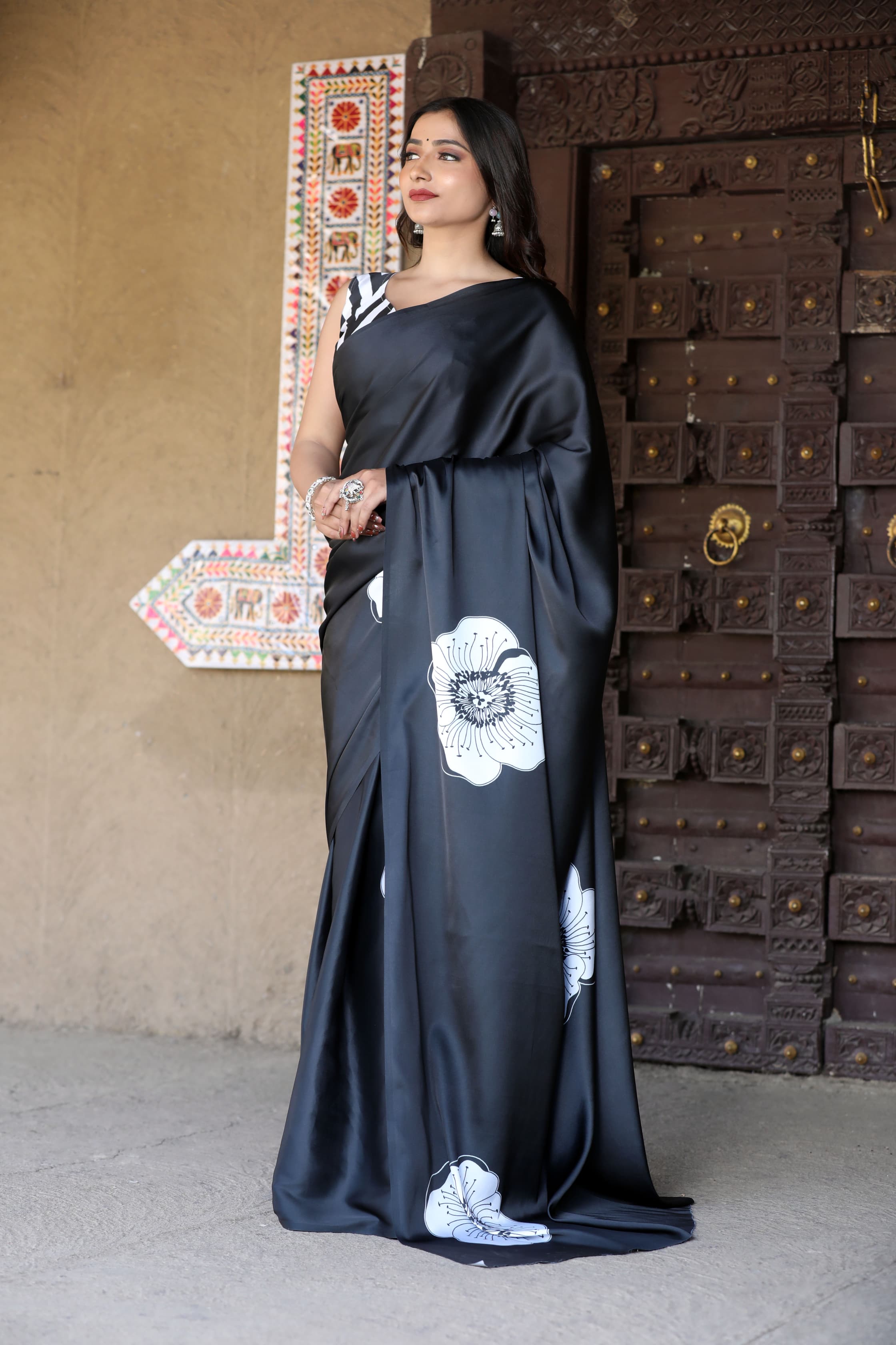Woman in a black saree with white floral patterns standing in front of a textured wall and wooden door.