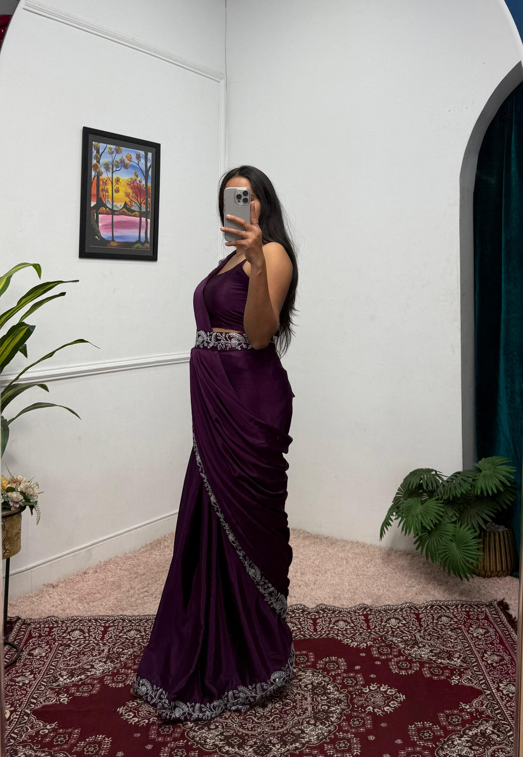 Woman in a purple evening gown taking a mirror selfie in a room with plants and a colorful painting.