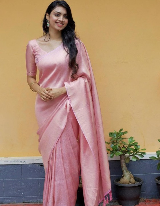 Woman in a pink saree standing against a yellow wall with a plant in the background