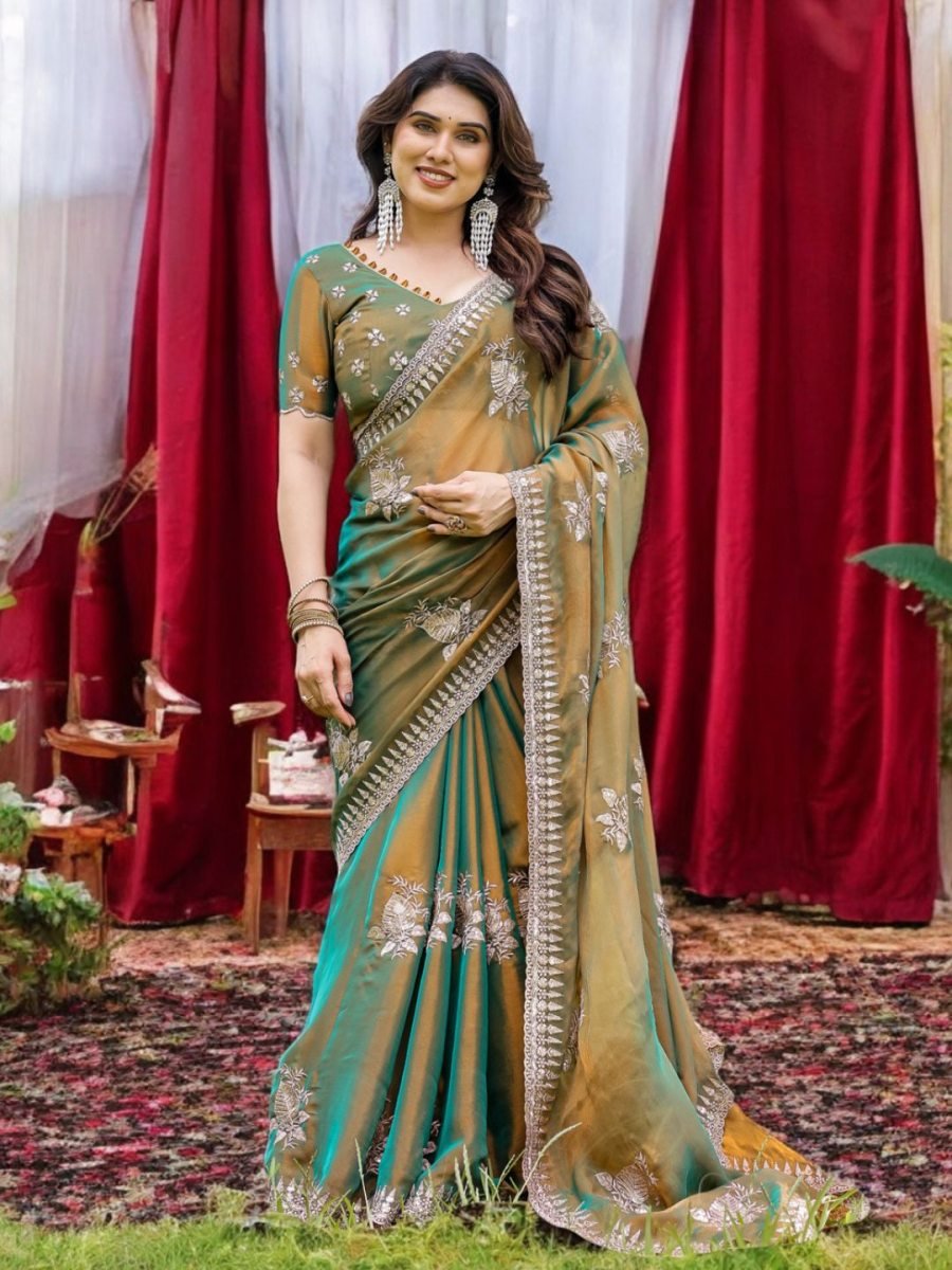Woman in a green and gold saree standing in front of red curtains.